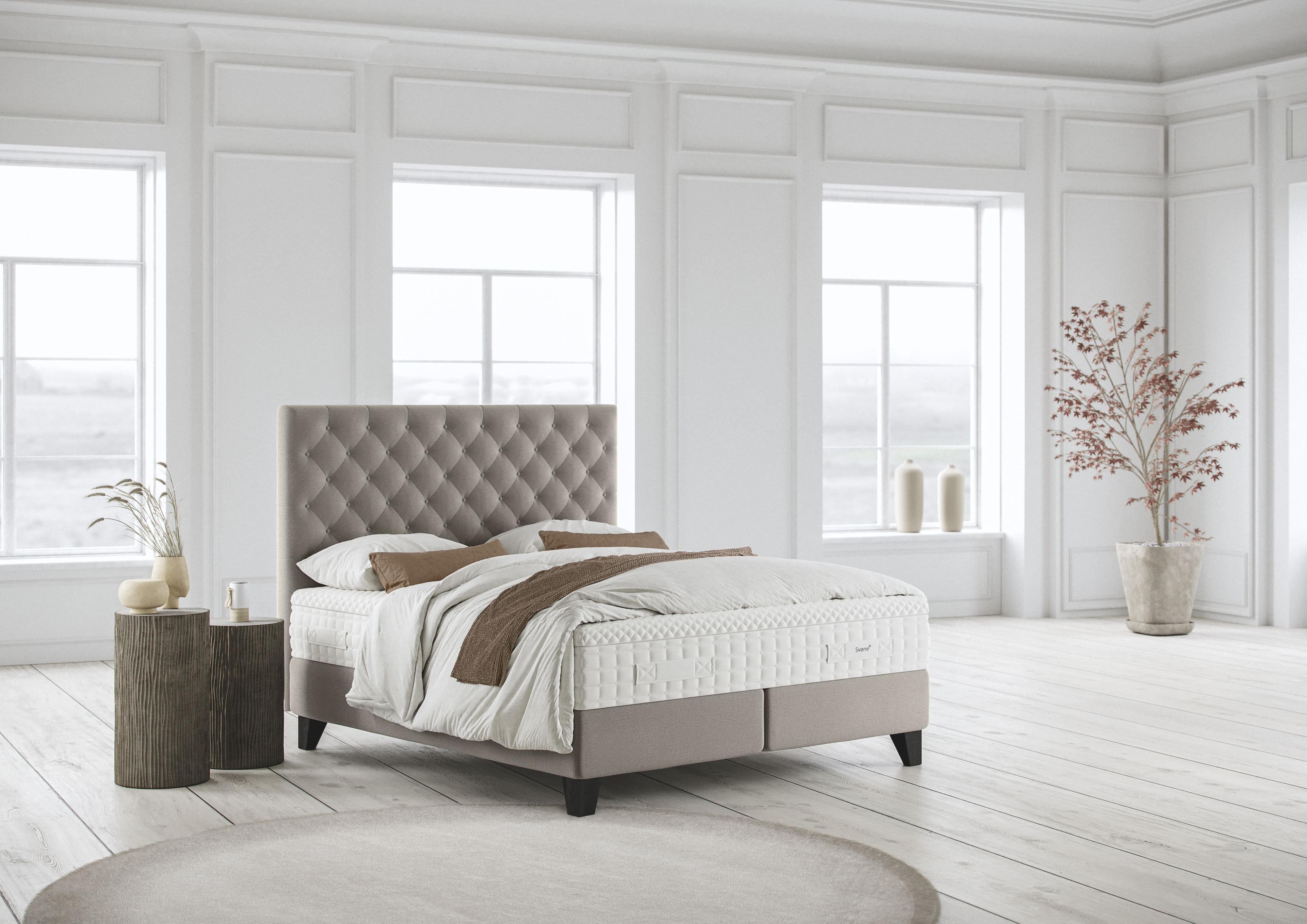 Schlichtes Schlafzimmer mit Svane Zensation Continental Bett in hellgrauem Stoff, gepolstertem Kopfteil mit Rautenmuster, zwei Nachttischen, großen Fenstern und hellem Holzboden.