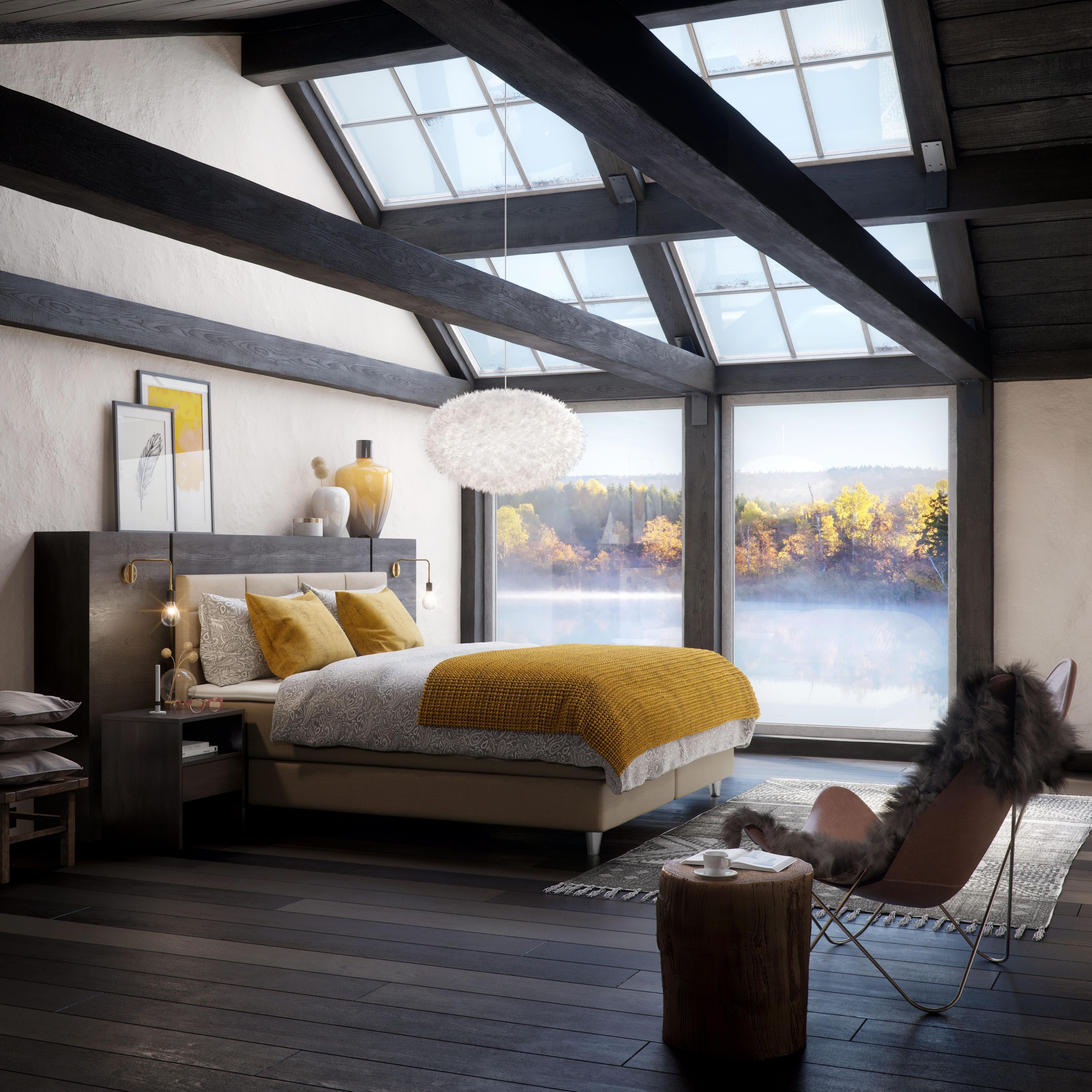 Schlafzimmer mit Svane Gelbett unter sichtbaren Holzbalken, großen Fenstern mit Blick auf herbstliche Landschaft, gelben Kissen und Decke, hängender weißer Lampe und Ledersessel mit Fellüberwurf.