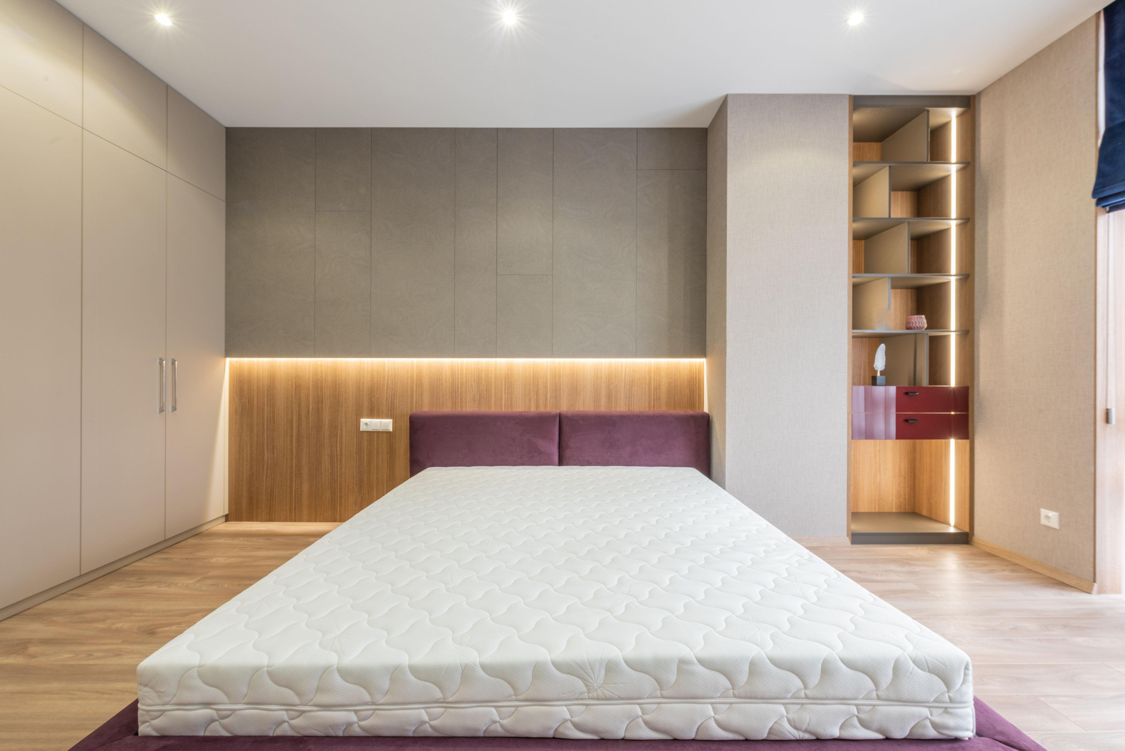 Schlafzimmer mit einer weißen Matratze auf einem lila Bettgestell vor einer Wand mit eingebauten Schränken und einem offenen Regal mit Beleuchtung.